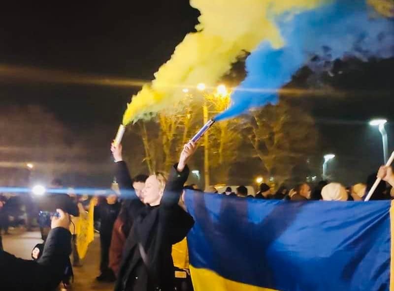 ID: Hand with index and middle finger raised in peace sign, overlaid with colors of the Ukraine flag, blue on top half, yellow on bottom.